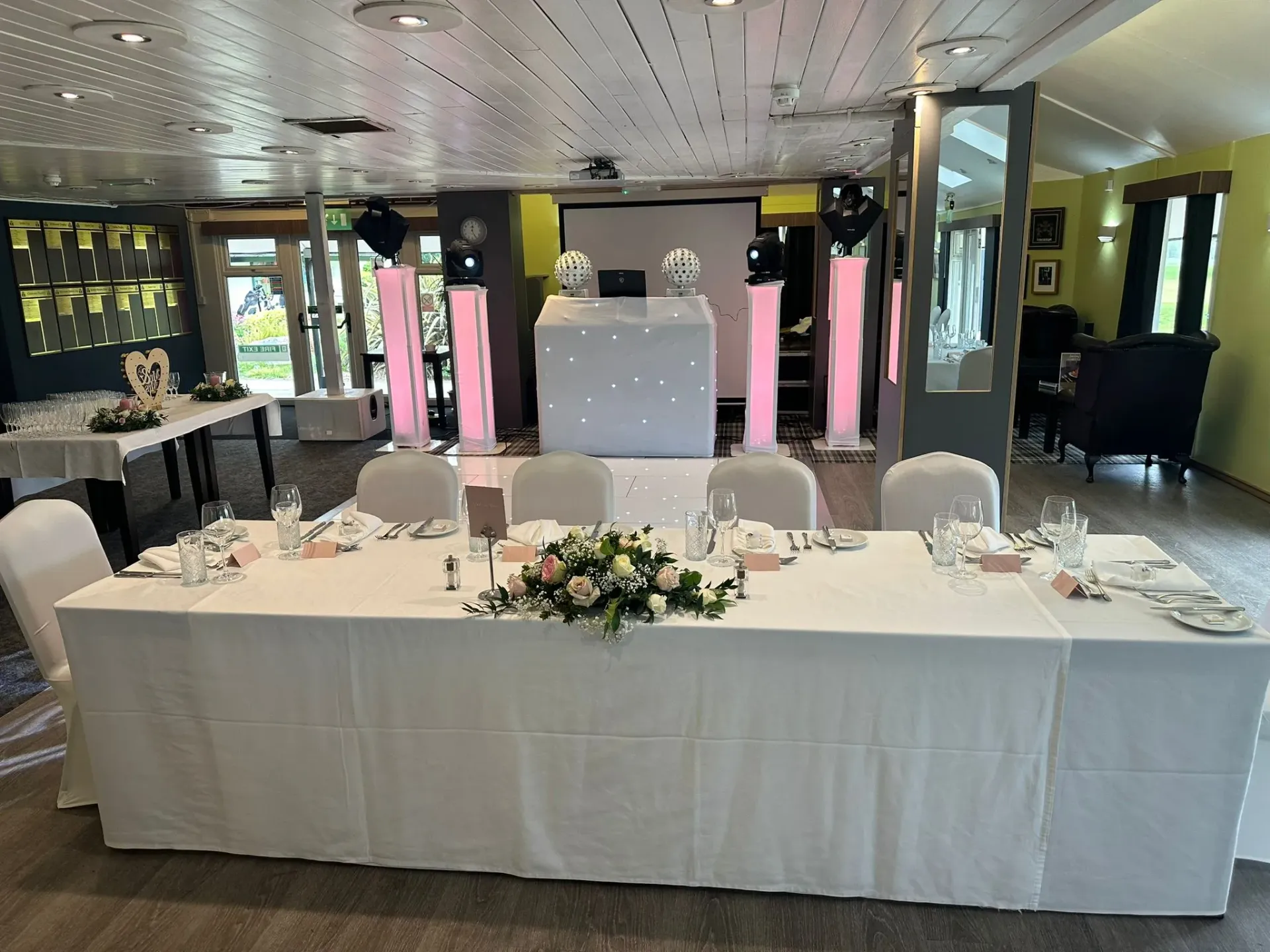 A stylish wedding reception room setup with a sparkling white DJ booth, pink uplighting on pillars, a large screen, and a reflective white starlit dance floor.