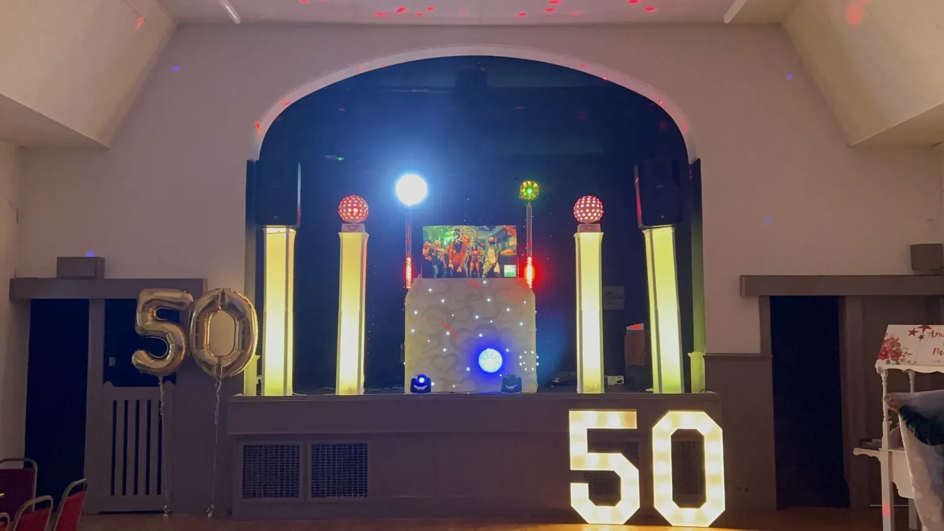 DJ setup with lights, screen, and large illuminated '50' numbers at a 50th birthday party.