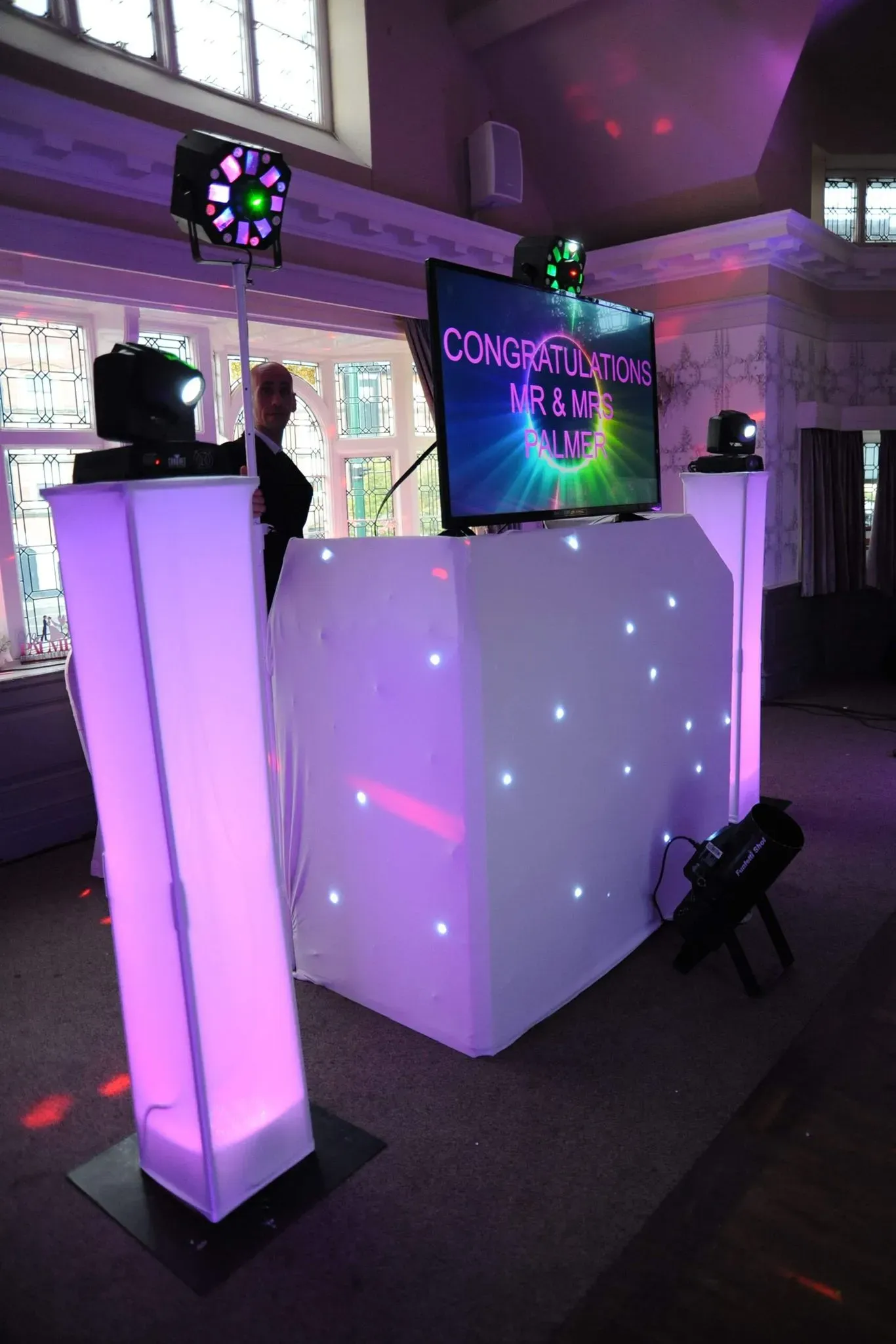 Award-winning Disco Entertainers DJ setup at a wedding reception, featuring a white LED DJ booth, purple uplighting, and a large screen displaying 'Congratulations Mr & Mrs Palmer' with a professional DJ visible.